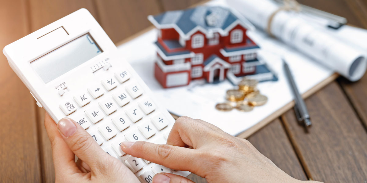 Close-up picture of a woman's hand operating a calculator and a model house in background - Using a Home Equity Loan for an Airbnb - DG Pinnacle Commercial - Miami Mortgage Lender