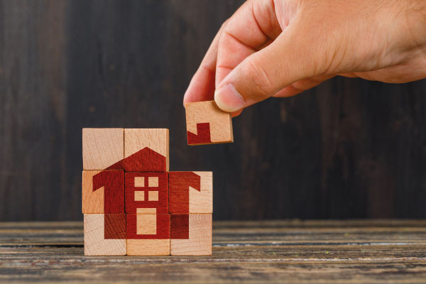 Hand setting up some wooden puzzle pieces with the printed figure of a house - 6 Benefits Of Investing In Real Estate - DG Pinnacle Commercial - Miami Real Estate Investor